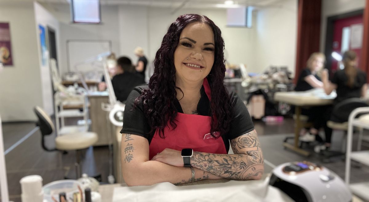 Woman with dark hair, wearing black top and pink apron sitting at nail table in training salon